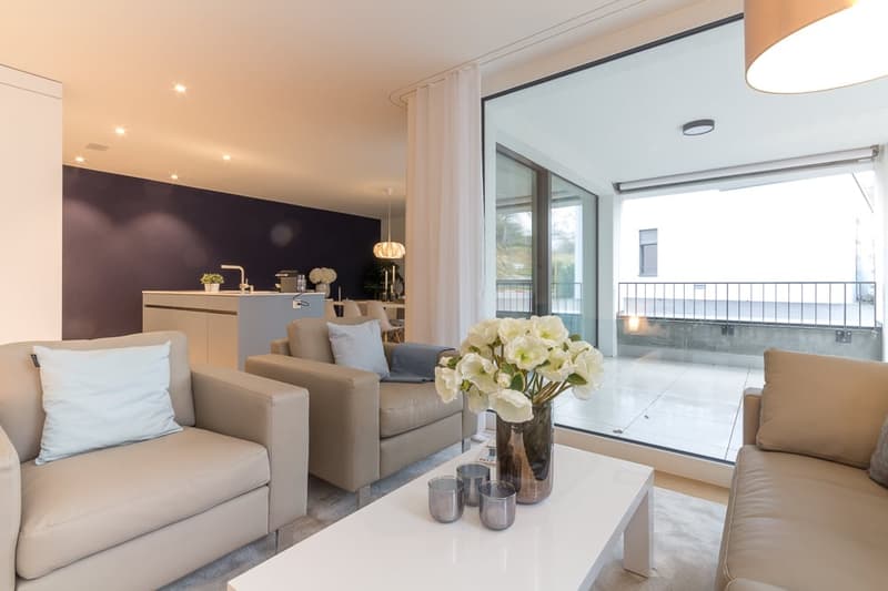 Beige leather couches, light wooden center table, large white flowers in glass vase, dark blue wall, large windows leading to balcony