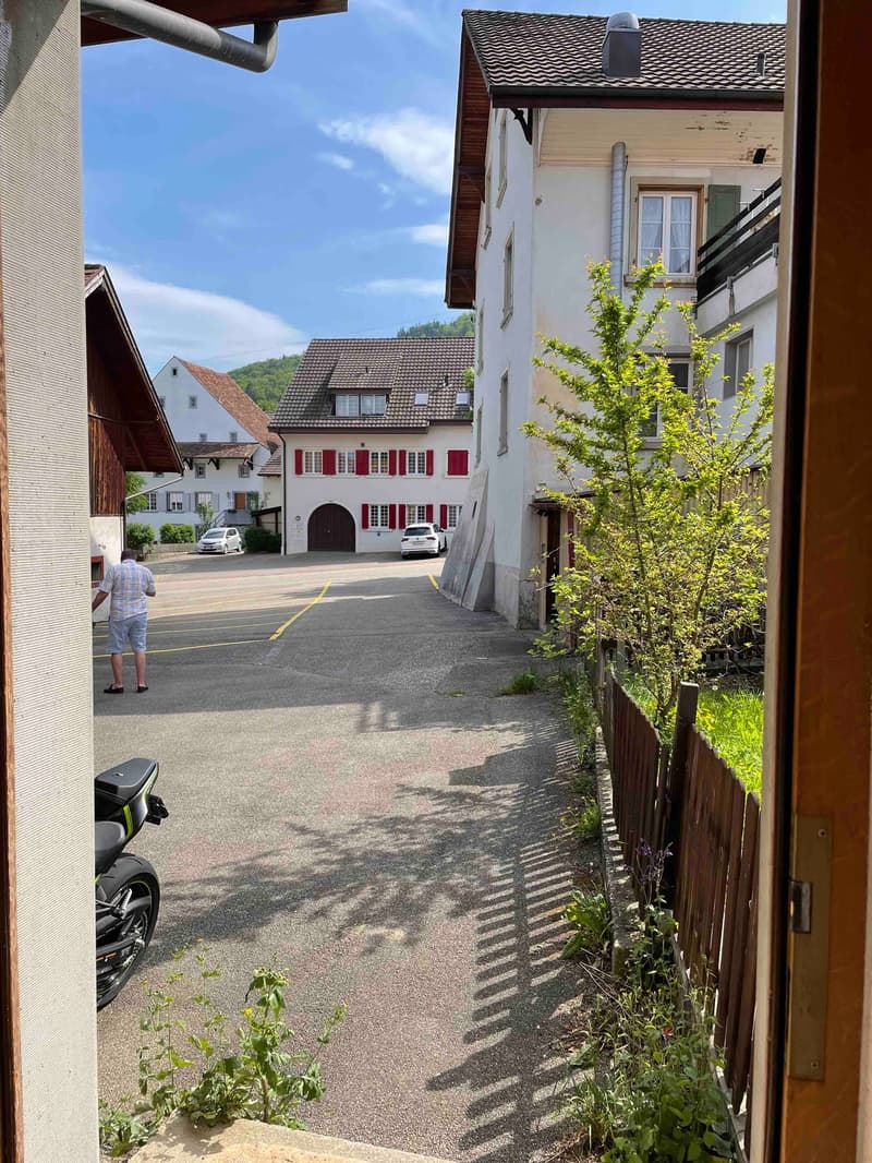 residential area, empty parking lot, man standing, building with brown roof, white building with red shutters, trees, greenery, mountains in the distance