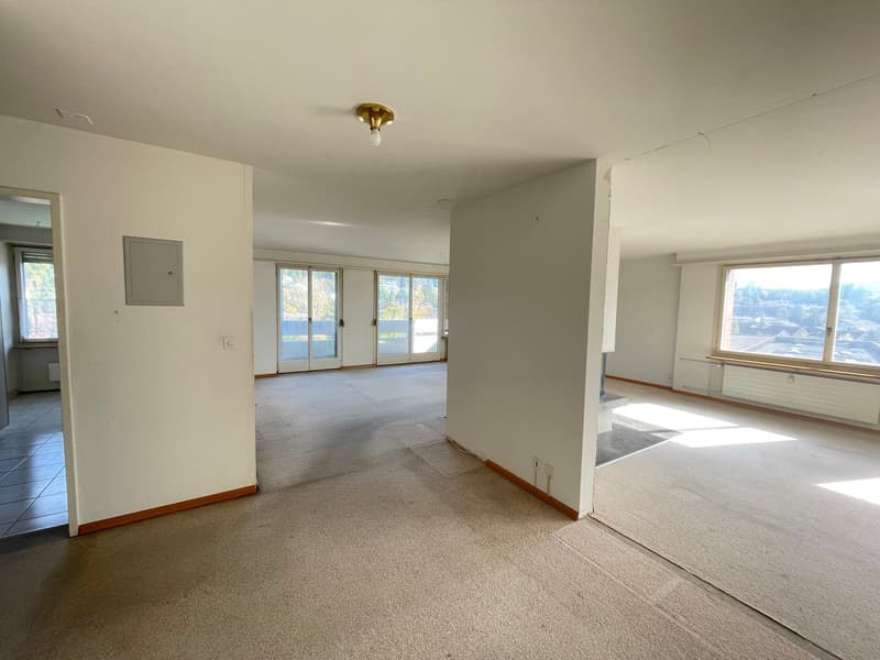 Empty room with white walls, carpet floor, ceiling lamp, windows with sliding doors, and a hallway leading to another room.