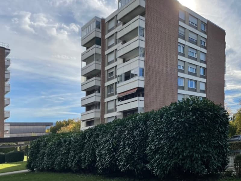 A multi-story brick building with many balconies and windows, surrounded by greenery and hedges.