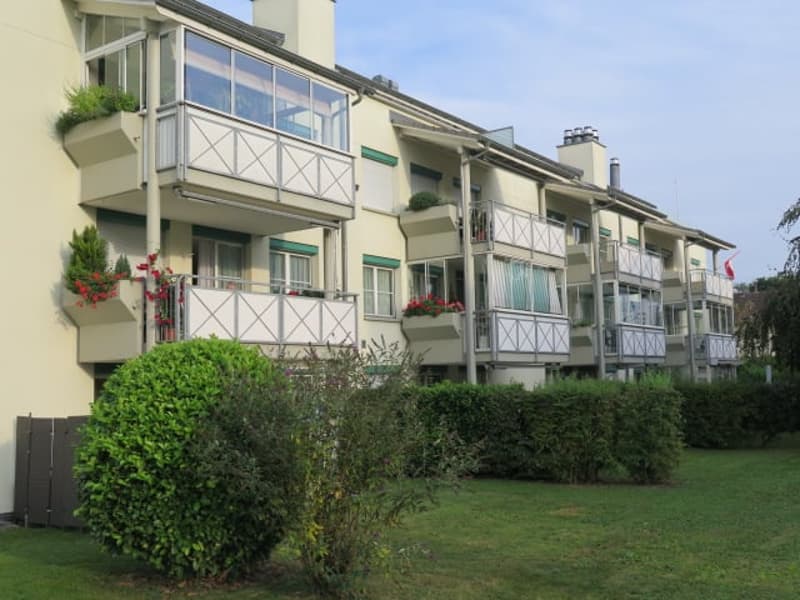 Multi-story apartment building with balconies, green landscaping, and a grassy area in the foreground.