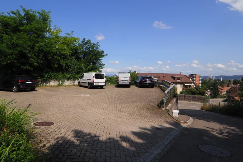 paved parking area with 4 cars parked, surrounded by trees and plants, with a view of the city