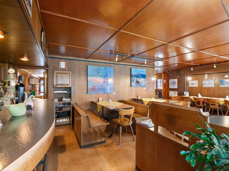 The image shows the interior of a cozy cafe or restaurant. The space features wooden paneled walls and ceilings, with track lighting overhead. There are several wooden tables and benches for seating, as well as a small counter area with what appears to be