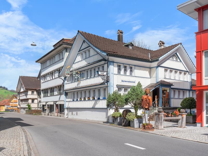 A traditional Swiss-style building with a red-tiled roof, white walls, and large windows. The building has a restaurant called 'Speiserestaurant' on the ground floor. There are some decorative elements like flower pots and sculptures outside the building.