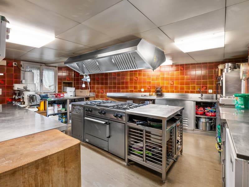 Large commercial kitchen with stainless steel appliances, gas stove, oven, dishwasher, and washing machine. The walls are covered in orange and yellow tiles, creating a warm and vibrant atmosphere. The kitchen appears to be well-equipped for commercial or