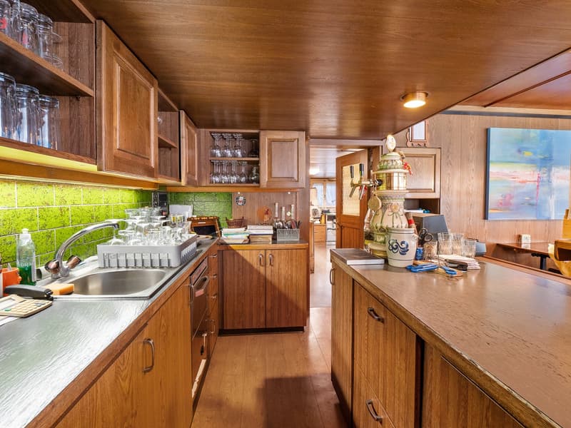 Spacious kitchen with wooden cabinets, green tile backsplash, stainless steel sink, and various kitchen appliances and utensils. The room has a warm, rustic feel with the wooden ceiling and walls.