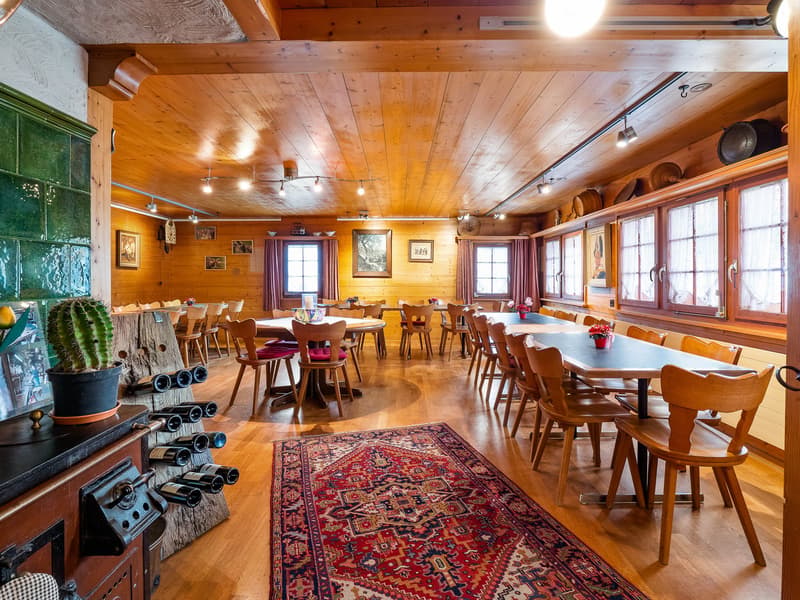 Rustic wooden interior with exposed beams, wooden tables and chairs, a large area rug, and a fireplace. The space appears to be a dining or restaurant area with multiple seating areas and windows providing natural light.