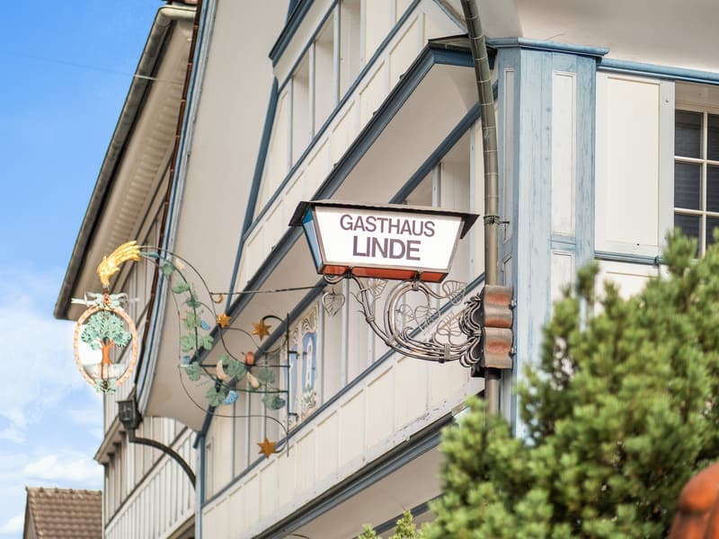 A traditional German-style building with a wooden facade, featuring a sign that reads 'Gasthaus Linde', indicating it is a restaurant or inn. The building has decorative elements like a wrought-iron sign holder and a clock on the exterior.