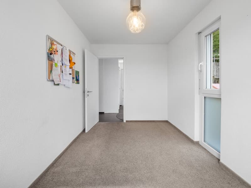 Empty hallway with carpeted floor, bulletin board, open door, light bulb hanging from the ceiling.