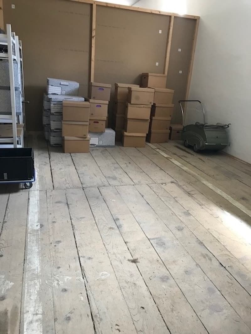 The image shows a storage room or garage filled with various boxes and equipment. The room has a wooden floor and appears to be used for storing items during a move or relocation.