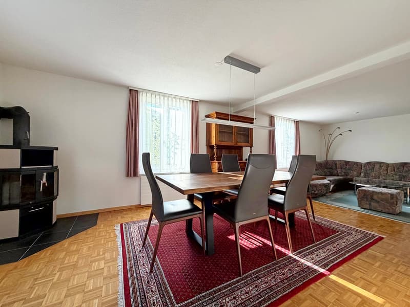 Dining room with a wooden table and chairs, rug, wooden flooring, large windows with curtains, cabinet, fireplace, lamp, couch, ottoman, and flooring
