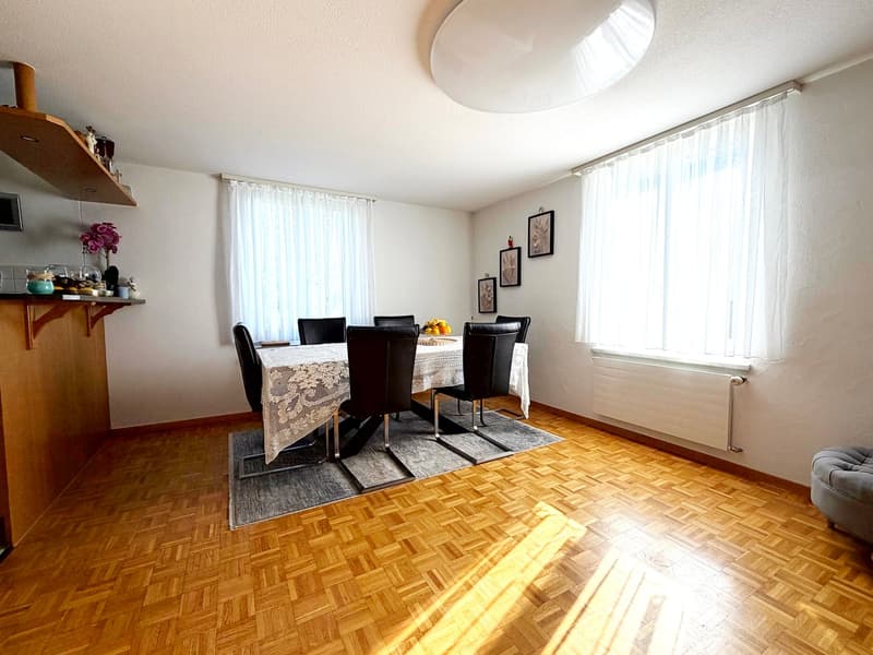 Dining room: Wooden floor, 6 chairs, table, white walls, ceiling lamp, windows with white curtains, shelves, fruit plate