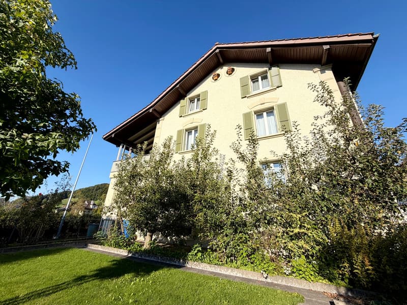 3 story yellow house, brown roof, green windows, green hedges, trees, balcony, grassy yard