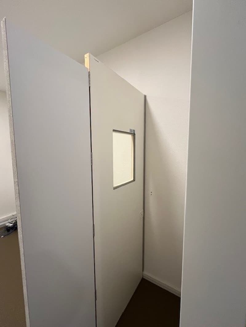 A corner with white walls and ceiling, an open white door with a small window, an exposed wooden beam, and a corner floor