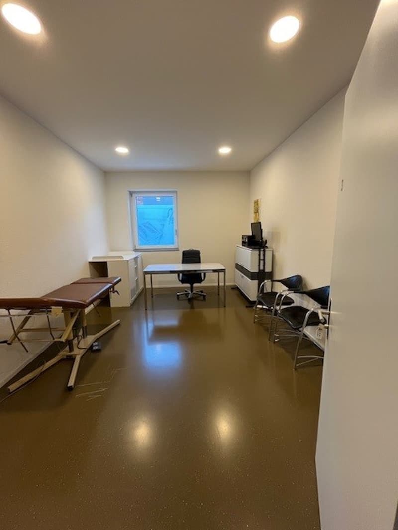 A room with several chairs, an office desk, and examination table. All of the furniture is brown in color, with a grey flooring. There is a window with a view outside.