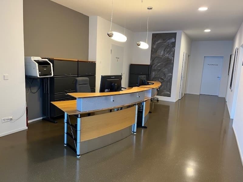Empty office space with modern desk, computer monitors, and office chairs. Equipped with a printer and cabinets. Bright white lighting.