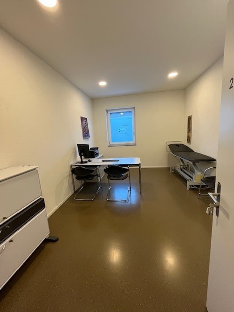 Office room with desk, chair, and window. Another chair and monitor are positioned against the wall. A medical examination table and cabinet are near the entrance. White walls and floors.