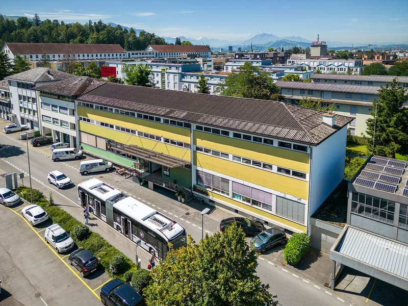 Large building with several stories, several cars parked in front, bus in front of building, solar panels on top of nearby building