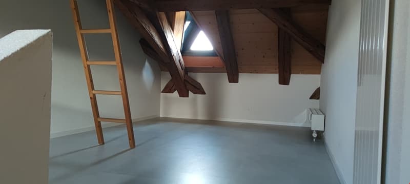 Spacious attic room with exposed wooden beams, triangular window, and ladder leading up to the space.