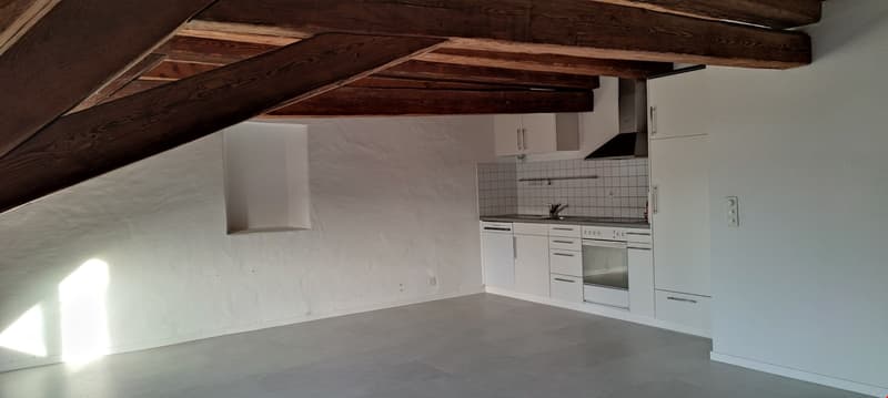 Attic apartment with exposed wooden beams, white walls, tiled kitchen with appliances, and concrete flooring.