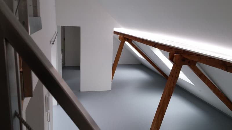 The image shows a room with white walls and a wooden table or desk with a triangular support structure. There is a white cabinet or shelving unit visible on the left side of the room. The room appears to be in a minimalist or modern style.