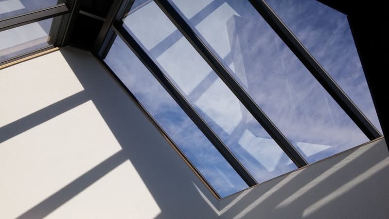 The image shows the exterior of a modern building with large, angular glass windows that provide a clear view of the blue sky and clouds outside. The windows are framed by sleek, black metal structures, creating a striking geometric design. The building a