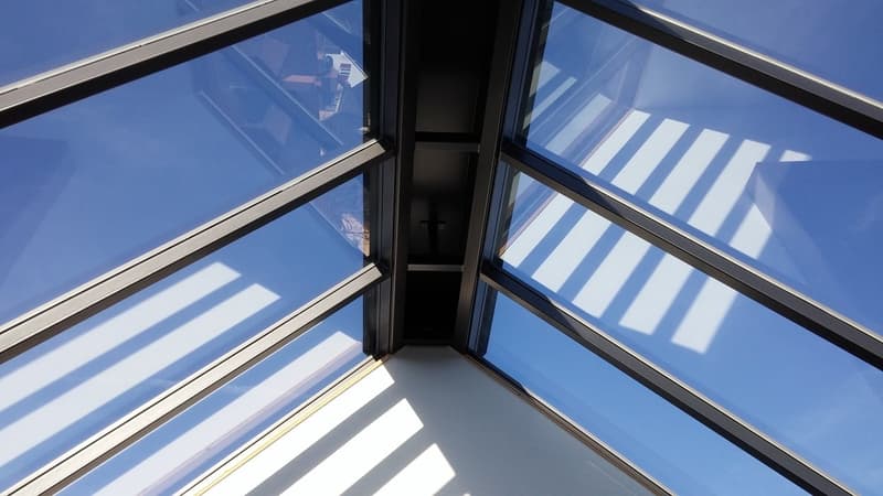 The image shows a glass structure with a slanted roof and large windows. The structure appears to be a skylight or atrium, allowing natural light to flood the interior space.