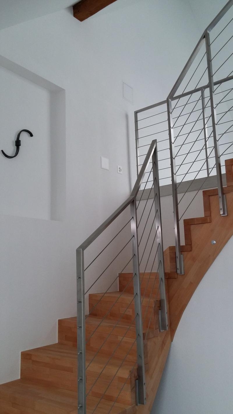 The image shows a modern staircase with a metal railing and wooden steps. The staircase appears to be located in a residential property, with a white wall and a wooden beam visible in the background.