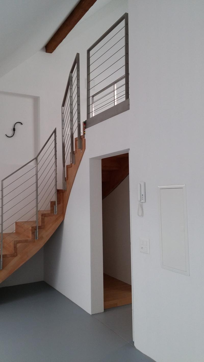 The image shows the interior of a building, with a wooden staircase and a metal railing system. The walls are painted white, and there is a wooden beam visible on the ceiling. The floor appears to be a dark-colored tile or concrete. There is a doorway vis