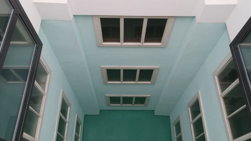 The image shows the interior of a building with a high ceiling and large windows. The walls are painted in a light blue-green color, and there are multiple rows of windows along the ceiling. The floor appears to be covered in a green carpet or tile. The o