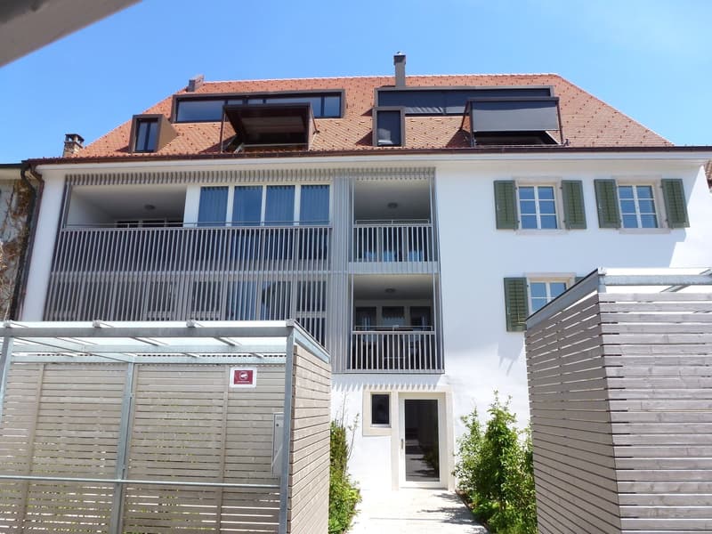 White building, red roof, 2 balconies, windows with shutters, entrance door, entrance path, metal fences on both sides