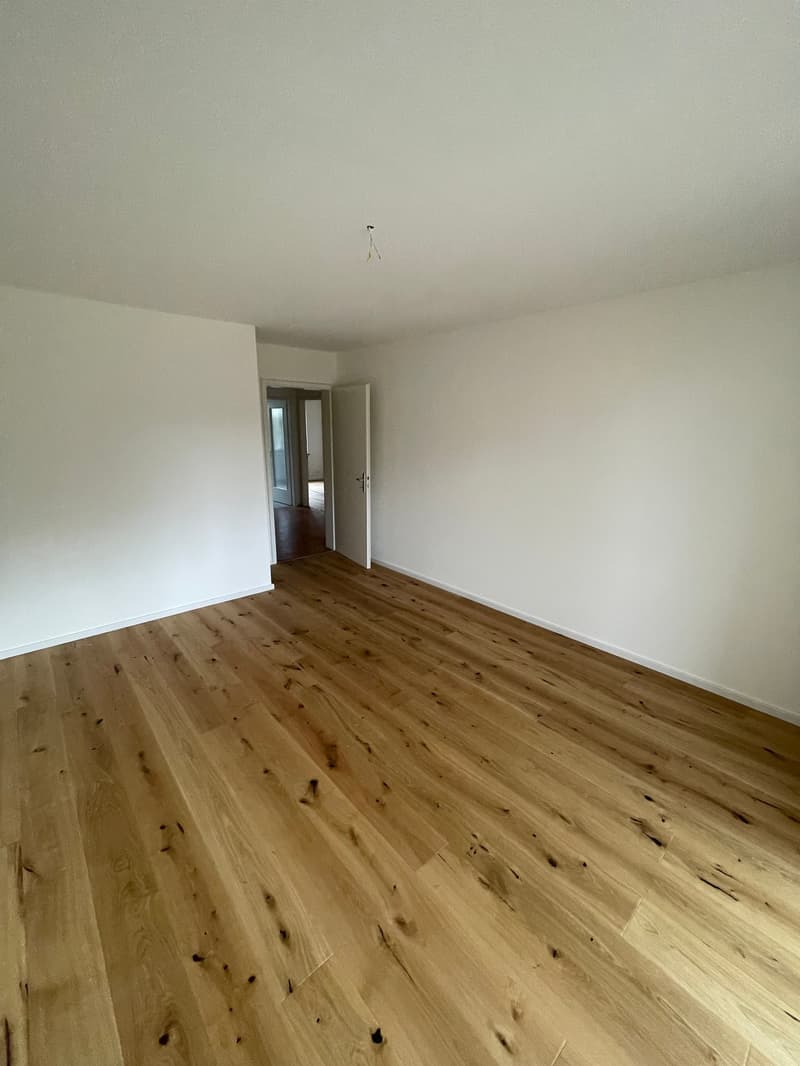 Large open room with hardwood floors, white walls, open door leading to another room.