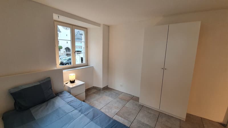 bedroom with bed, nightstand with lamp, wardrobe, window with outdoor view, beige walls, tiled floor