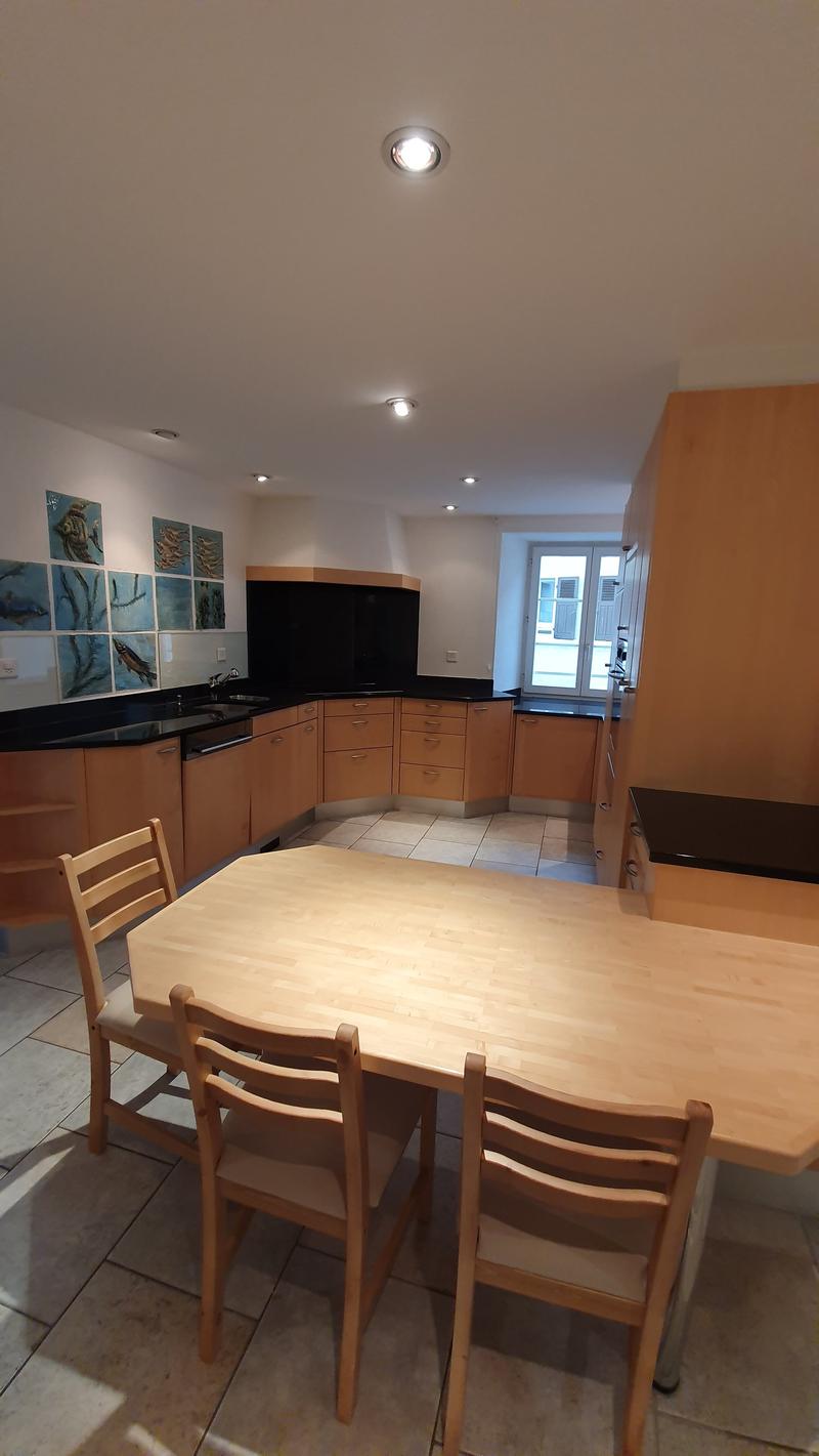open kitchen with wooden cabinets, black countertops, wooden dining table with chairs, tiled floor, wall-mounted ceiling lights, central light, artwork on the wall, sink with faucet, dishwasher, large window