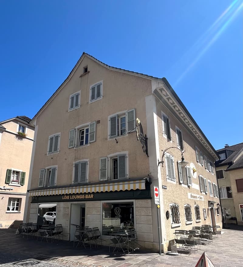 Two-story building, beige stone facade, many windows with white shutters, a cafe with a terrace on the ground floor, awning on the cafe, outdoor tables, reflection of a white car in the window, cobblestone street.