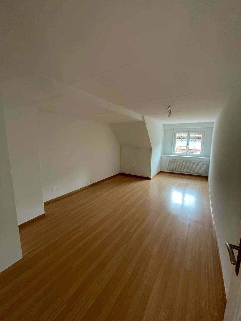 This image shows a spacious room with hardwood flooring, white walls, and a large window. The room appears to be empty, with no furniture or decor visible. The room has a clean, minimalist aesthetic.