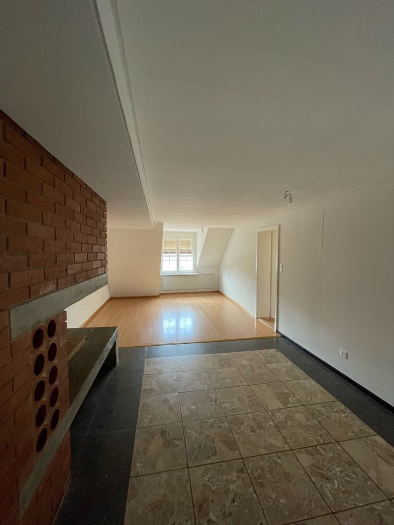 The image shows an empty room with a brick wall on one side and a tiled floor. The room has a hardwood floor and white walls, and there is a window visible. The room appears to be a living or dining area.