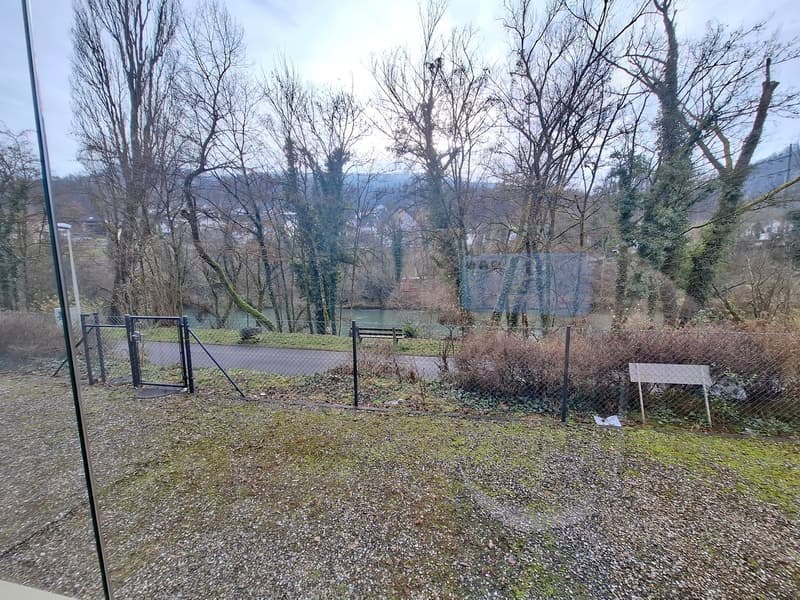 A gravelled area with bare trees, a pathway, a bench by the water, and mountains in the background