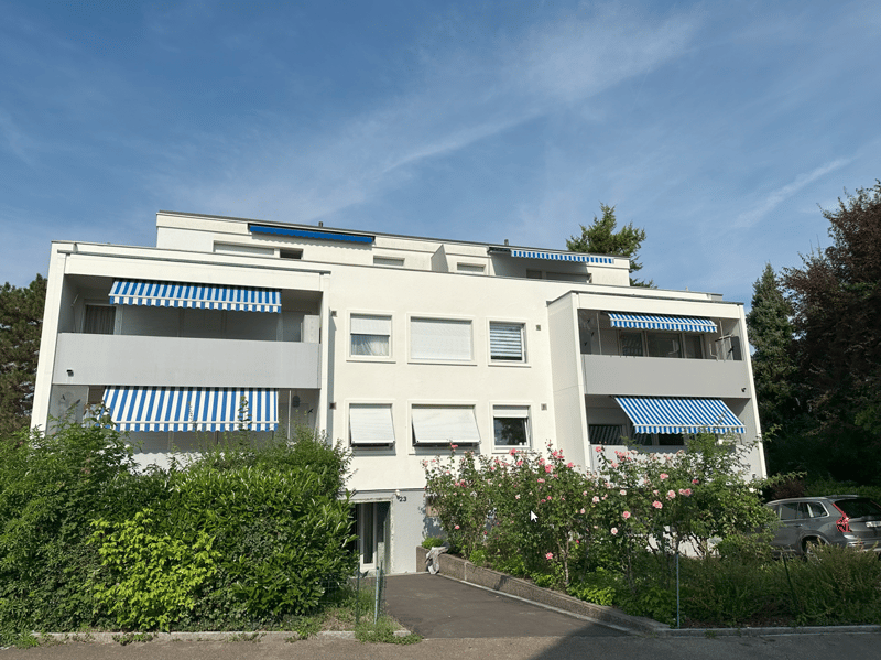 Modern white 3 story building, multiple balconies, with greenery, entrance, car parked