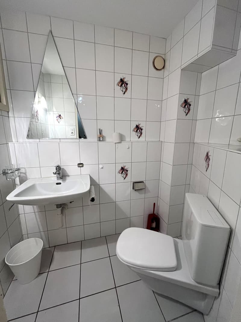 Bathroom with white tiles, sink, toilet, mirror, and a trash can