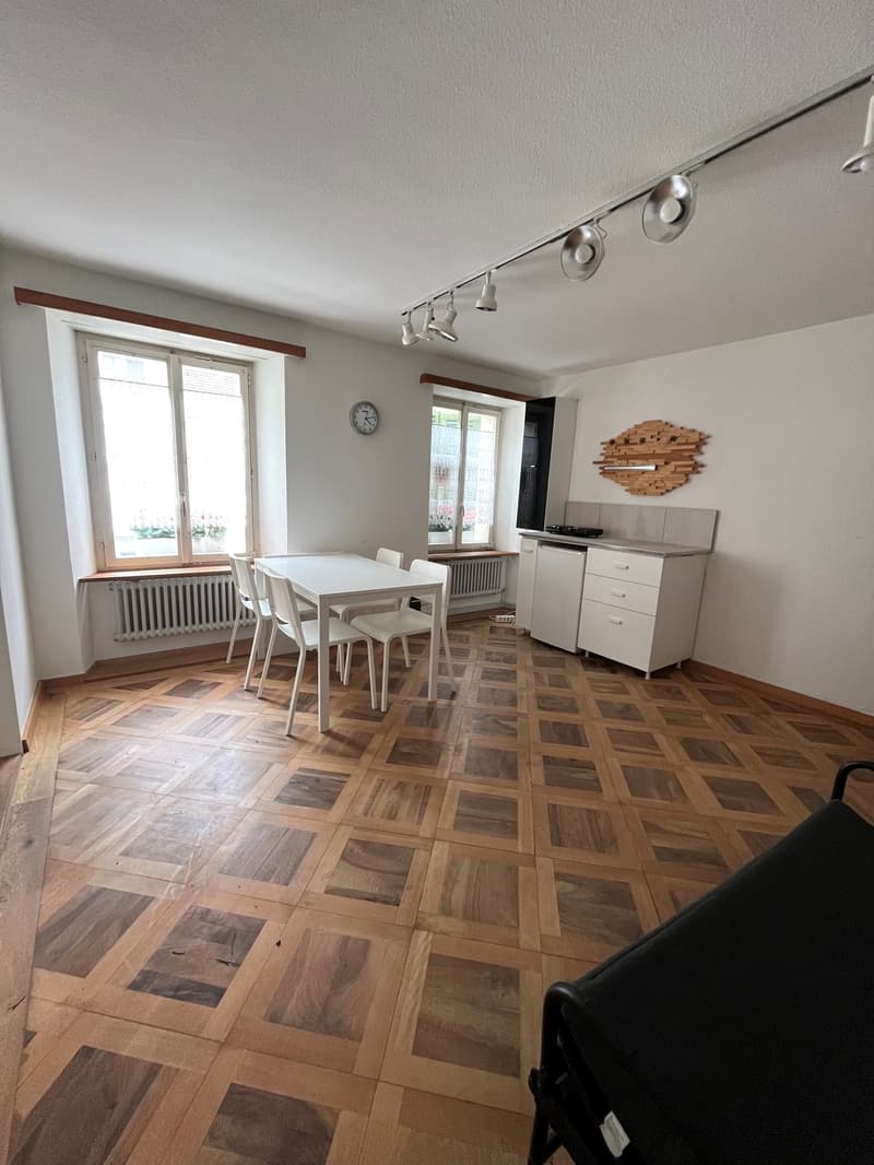 open dining area, parquet flooring, white walls, large window, dining table, four chairs, radiator, clock, kitchen area, kitchen cabinets, countertop, folding bed