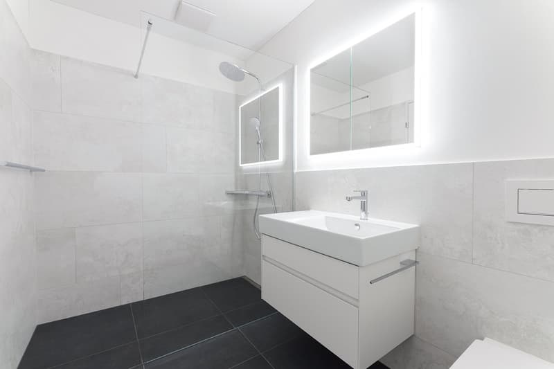 modern bathroom, white tiles, black tiles on floor, glass shower enclosure, white vanity with sink, mirror with lighting