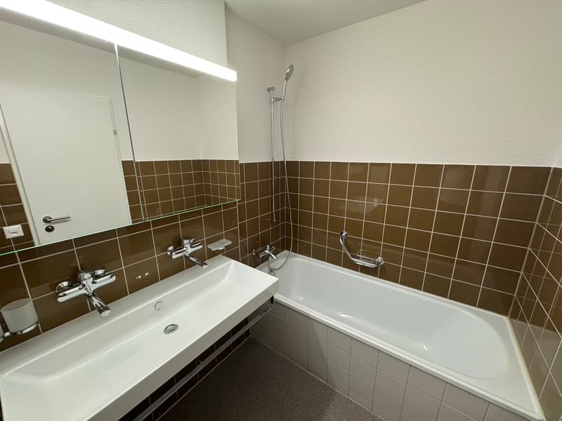 Bathroom with brown tiles, white bathtub, white sink, silver faucets, showerhead, mirror, and a power outlet