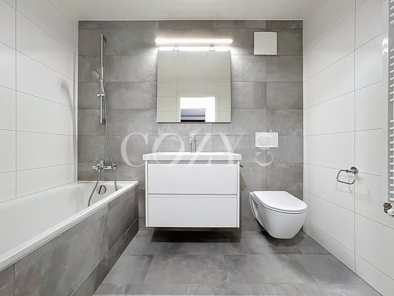 Large bathroom with grey tile walls and floors, white vanity with sink, toilet, bathtub, and shower with handheld showerhead.