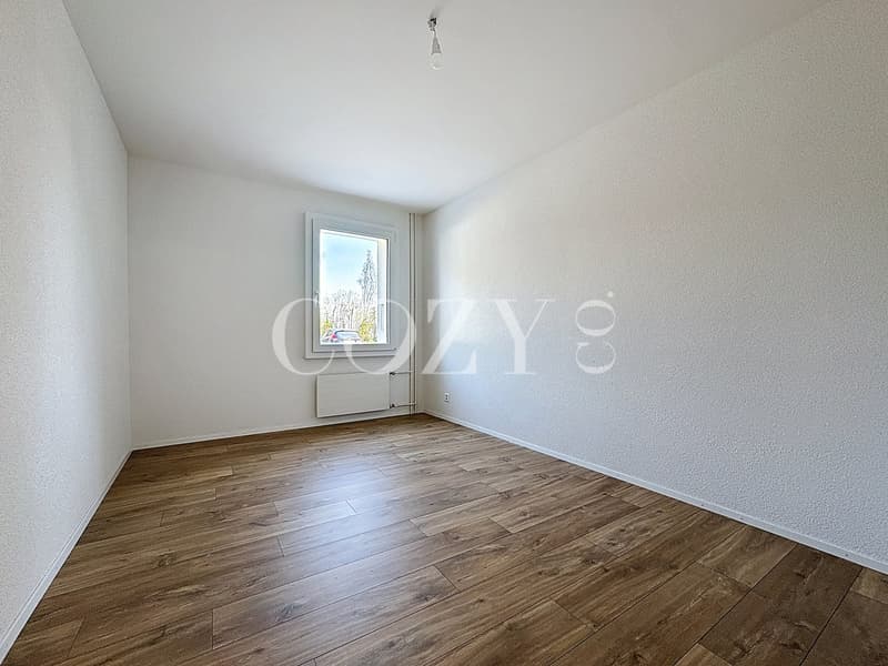 Spacious bedroom with white walls, hardwood flooring, and a window providing natural light.