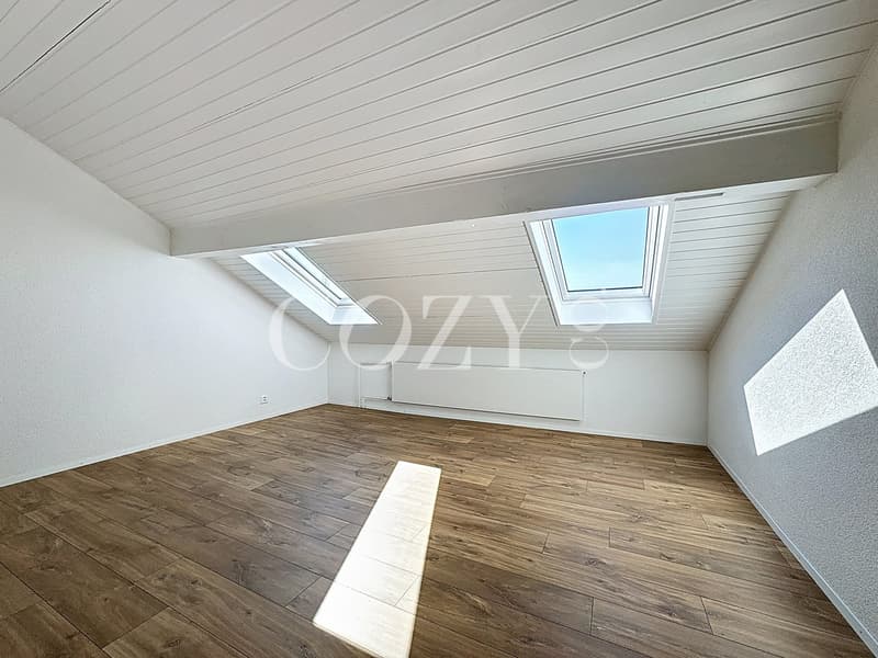 Spacious attic or loft space with white walls, a slanted white ceiling with wooden beams, and large skylights providing natural light. The floor is covered with wood-look laminate flooring.