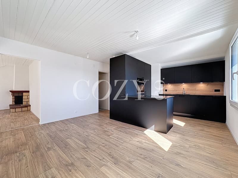 The image shows an open-plan living space with a modern kitchen featuring black cabinets and a wooden backsplash. The room has a high, white-paneled ceiling and hardwood flooring. A fireplace is visible in the corner of the room.