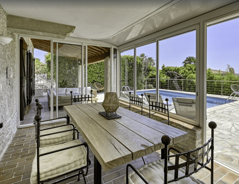 Outdoor dining area with wooden table and chairs, glass doors, patio with swimming pool view, couch, and garden.