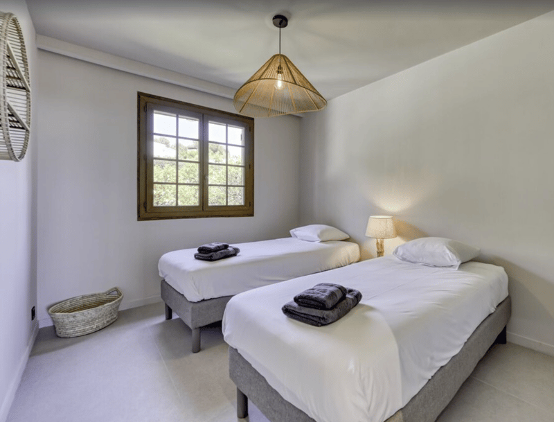 Bedroom with two single beds, white walls, wooden window frame, lamp, ceiling fan, white bedding and towels
