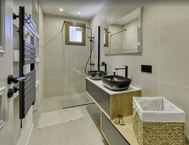 Bathroom with white tiles, shower, black basins, mirror, wooden counter, wicker basket.